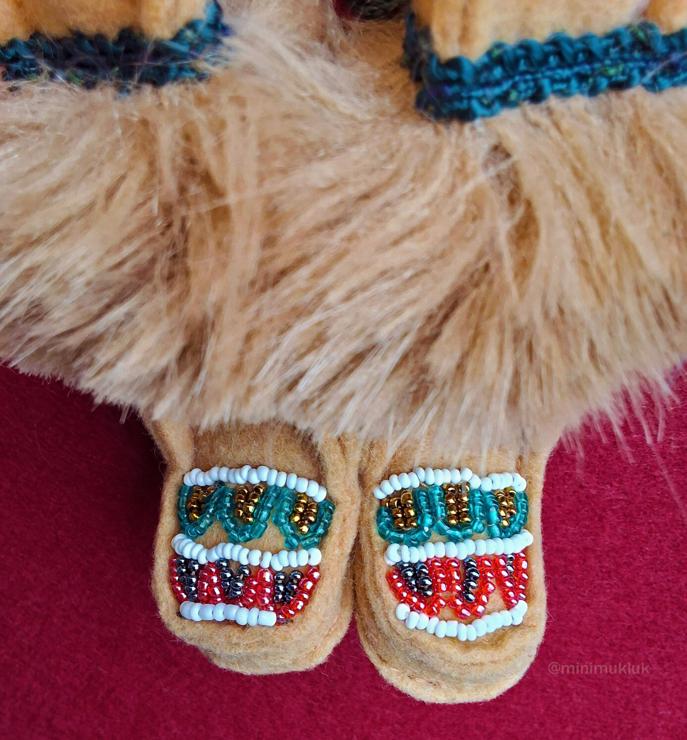 A pair of brown felt boots. Between white bead, a arrow pattern repeats. Teal green, gold, red, and purple seed beads are sewn in. A green trim with matching brown fur along the top.